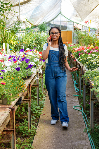 Joyful African-descent woman chats on phone surrounded by vibrant greenhouse flora in casual denim attire.