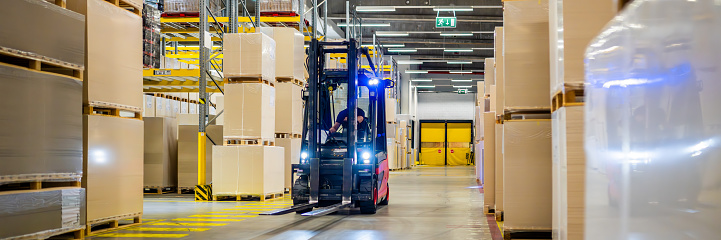 Warehouse worker driving forklift truck, illuminating cardboard boxes with headlights