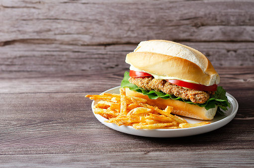 Milanesa sandwich with tomato slices, lettuce and mayonnaise, and French fries in a white plate on a wooden background with copy space.
