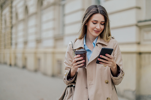 Beautiful young fashionable female walking in the city streets and using a mobile phone while drinking coffee takeaway. Stylish woman in trench coat typing message on a smartphone. Copy space.