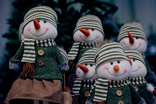 A group of snowmen with red carrot noses dressed in green sweaters, scarves and striped hats under the Christmas tree. Christmas decorations.