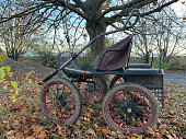 An old open single-seater four-wheeled horse-drawn carriage without a horse. A tarantass or carriage stands under a tree in autumn among fallen leaves in sunny weather in a German village in Germany.