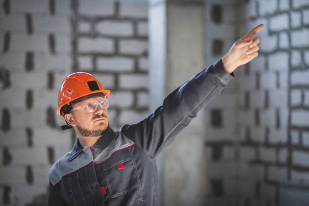 istock Bearded worker in blue uniform and helmet pointing with finger to the side at construction site 1809989478