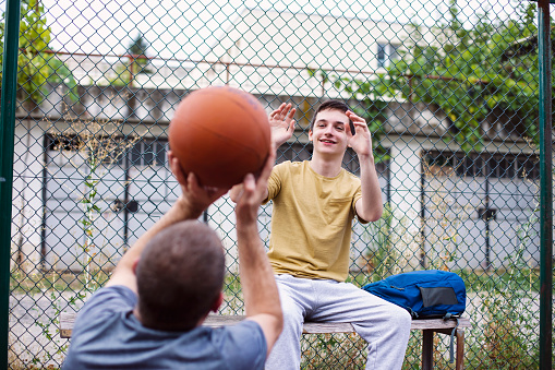Middle-aged man and teenage boy having fun and talking about basketball. Playing and learning sports game outdoor.
