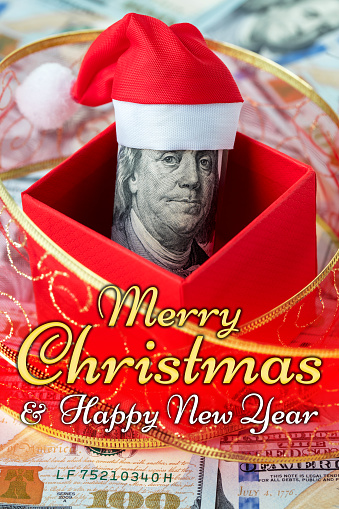 A gift box with a hundred dollar roll in a Christmas Santa hat as a Christmas present. US 100 dollar bills background. Merry Christmas and Happy New Year lettering.