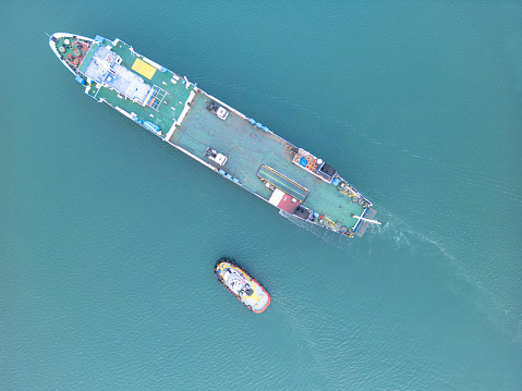 The tug accompanies the ship when entering the cargo port. \nAerial drone view above sea port. Cargo vessel is loading containers and bulk. Professional business logistics and transportation of cargo ship.