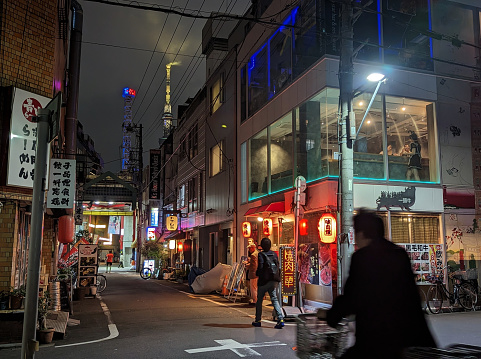 Taito City, Japan - May 22, 2023: Pedestrians and cyclists go along 2-Chome in the Asakusa District. The illuminated towers of Asakusa Hanayashiki  and Tokyo SkyTree in Sumida stand in the background.