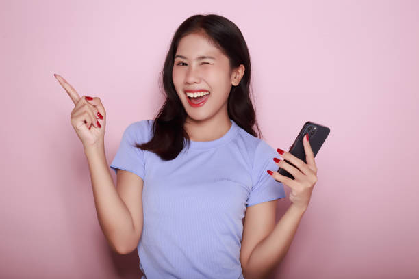 istock A young woman poses with her finger pointing in an open space while holding a phone in her other hand, Portrait of a beautiful young woman in a light pink background. 1810050178