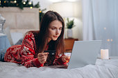 Christmas online shopping, winter holiday sales and discounts, online shopping at home. Woman lying on bed with laptop with credit card and Christmas decorations