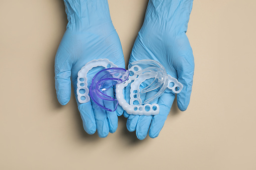 Bite correction. Dentist in medical gloves holding different mouth guards on beige background, top view