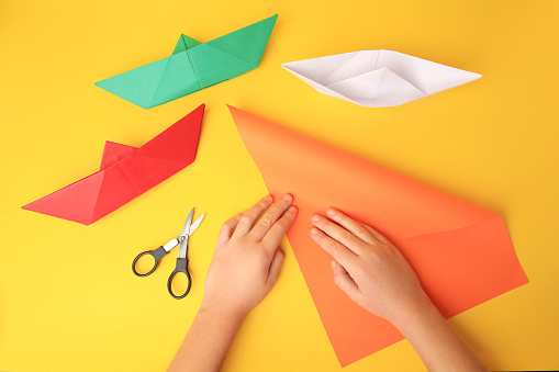 Origami art. Child folding paper on yellow background, closeup and top view