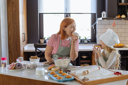 A cute girl with blond hair and her mom are wearing aprons and enjoying together in the kitchen