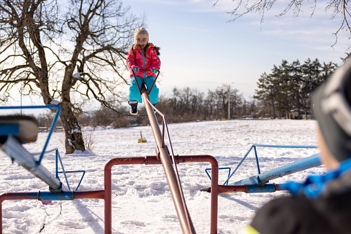 An over-the-shoulder view of a Caucasian  father playing on the seesaw with his toddler daughter, on the playground in nature, during a winter day