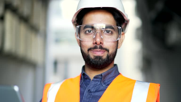 Close up portrait of young smiling professional engineer wearing safety helmet, protective glasses and vest standing at factory.