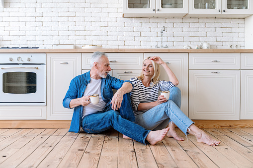 Celebrate moving to new home. Smiling mid aged woman and man sit on wooden floor in cozy kitchen, enjoy morning, drink coffee together. Mature couple making plans on future
