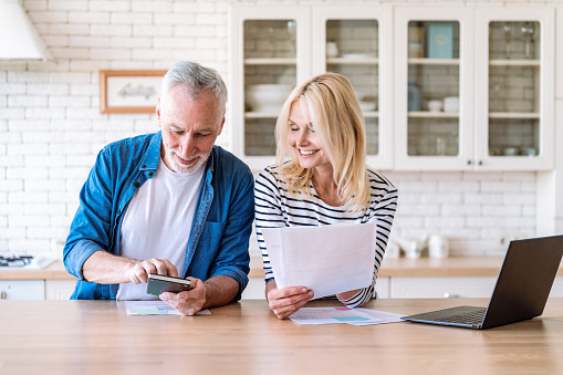 Happy mature family couple managing household budget, planning investment, analyzing paper utility bills, making payments and fill application forms together