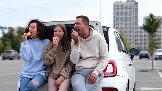 Caucasian joyful nice family woman and man with teenager girl smiling doing cheers gesture with donuts in hands. Parents with teen daughter eating tasty doughnuts enjoying donut sitting in car trunk