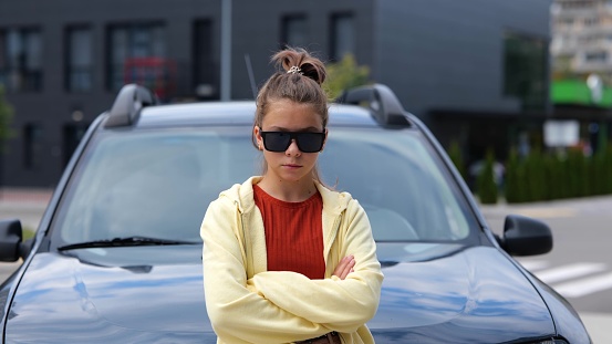 Pretty Caucasian cool young teenager girl in black sunglasses standing in front of car on street looking at camera chewing a gum. Close up. Stylish pretentious teen smiling at camera. Slow motion