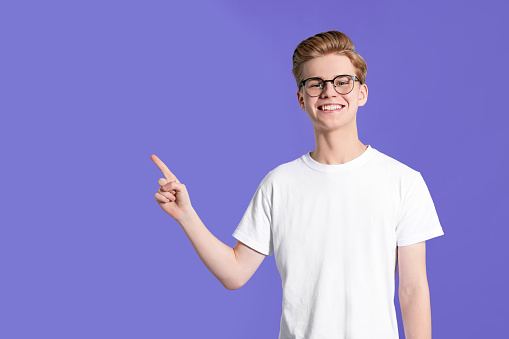 Teenage boy pointing at something on purple background. Space for text