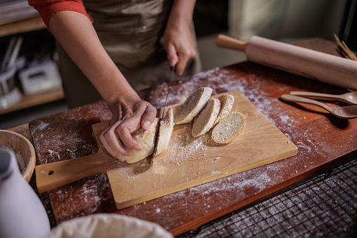 Close up of a baker's hands as they slice freshly baked bread on a rustic wooden board, with a dusting of flour and baking utensils around, evoking warmth and homeliness