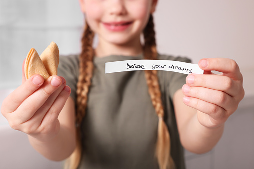 Girl holding tasty fortune cookie and prediction indoors, closeup