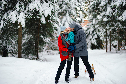 Happy couple outdoors on the snow with their cute little daughter. They are holding her between them and kissing her on the cheeks.