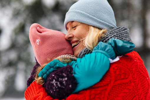 Close up shot of a beautiful woman wearing a winter sweater and a hat, hugging her child while enjoying a winter day outside.