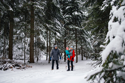 Smiling couple with their cute little daughter, all dressed in winter clothes, walking through the forest covered in snow. Father is pulling the sleds while the mother is carrying their child.