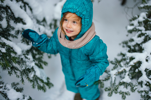 Close up shot of an happy adorable little girl in winter clothes playing and shaking snow of trees on a nice day outside. She is looking away and smiling.