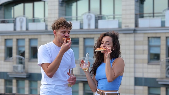 Portrait of young carefree couple male and female dancing while drinking beer and eating pizza at a party. Joyful casual people having fun at party on a roof. Fun. Good mood. Leisure concept