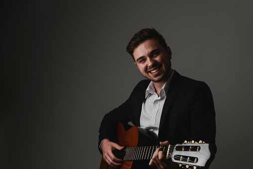 Handsome young man smiling and playing guitar while sitting against grey background