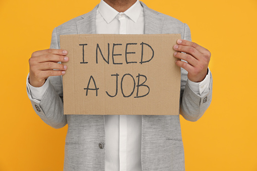 Young unemployed man holding sign with phrase I Need A job on yellow background, closeup