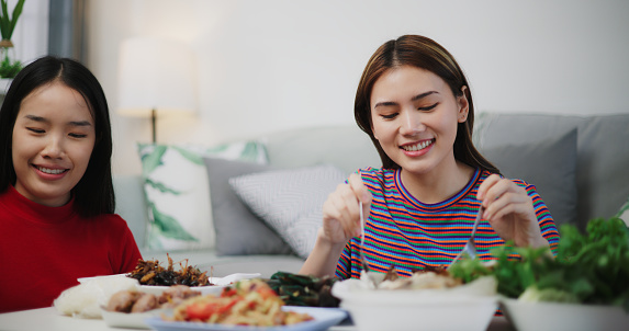 Image of Happy young Asian woman eating Thai food while sitting on the floor in the living room at home. Women friends fun joy relax enjoy a Thai meal. Lifestyle, People and Thai food concepts.
