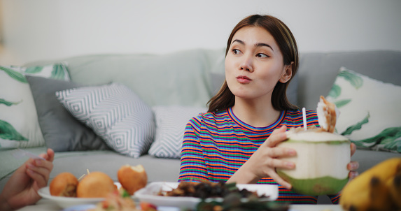 Image of Young Asian woman eating Thai food and drinking coconut juice while sitting on the floor at home. Women friends fun joy relax enjoy a Thai meal. Lifestyle, People and Thai food concepts.