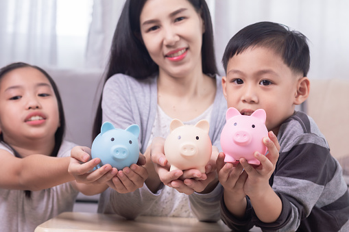 Asian family shows piggybank full of coins to camera, practicing finance planning for future, mother put coins in piggy bank and encourage children to be disciplined to save money left from school