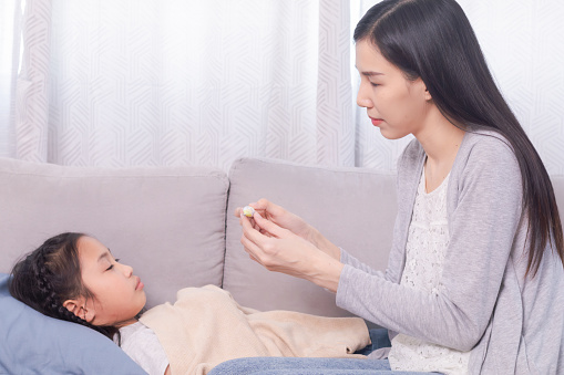 Asian woman using thermometer to check high-temperature girl sickness headache at home, mother watch children lying down on sofa while mom worried daughter get fever illness, medical healthcare