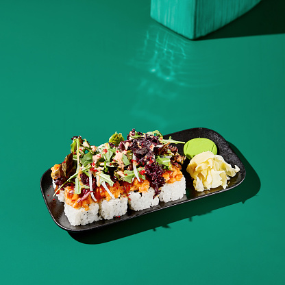 Artistic sushi roll with salmon tartar and salad mix on a black plate against a vivid green background. Modern minimalist composition for Asian restaurant menu.