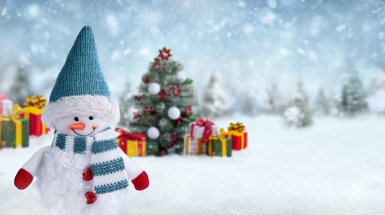 Christmas background with happy snowman dressed in red mittens, hat and scarf in winter landscape. Blurred Christmas tree and gift boxes on background. Gift card or banner with copy space.