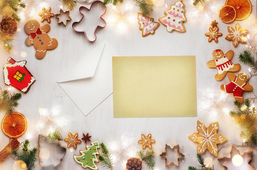 Envelope and blank letter to Santa Claus with a place for text on the background of Christmas gingerbread cookies, festive food decorations and garland with lights. Christmas and New Year holiday background. Xmas greeting card. Flat lay. Part of the series.