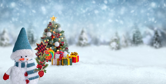 Winter or Christmas panoramic background with happy snowman holding a fir branch decorated with berries, candy and star shaped decoration. Blurred Christmas tree and gift boxes on background. Gift card or banner with a snowman dressed in red mittens, hat and scarf in winter landscape.