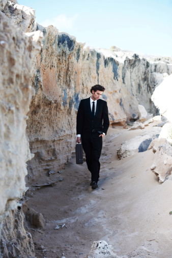 Handsome businessman strolling along the sand next to a rock formation, carrying a briefcase