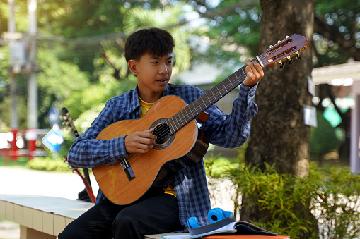 Asian boy happily sits outside playing guitar with his camp friends during break. Soft and selective focus.