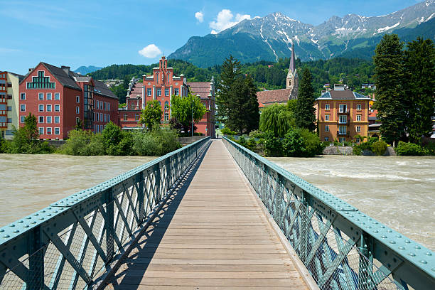 A pedestrian bridge spans the Sill River in Innsbruck, Austria. A person on a bicycle is approaching the bridge.