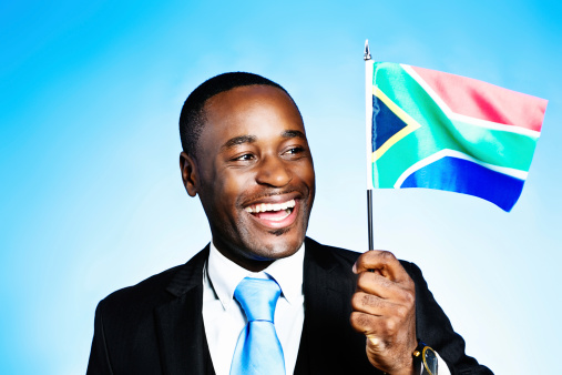 This patriotic, handsome  African businessman or politiican gives a beaming smile as he waves the South African flag against a blue sky.