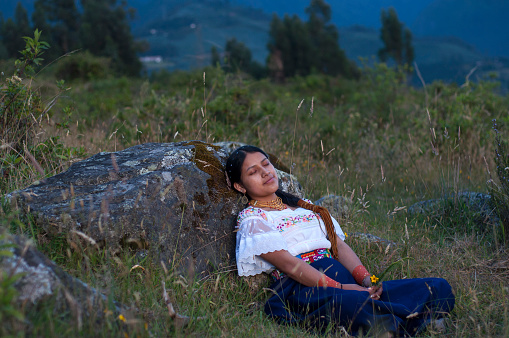 copy-space of a beautiful indigenous woman from ecuador relaxing on a rock in the mountain in a lush landscape. Hispanic heritage month. High quality photo