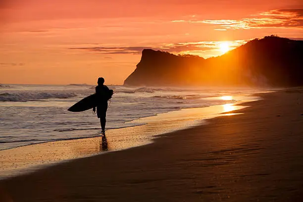 A silhouetted surfer walks down a beach in Nicaragua on sunset during a balmy June afternoon with mountains and ocean in the background. Lots of copy space. 