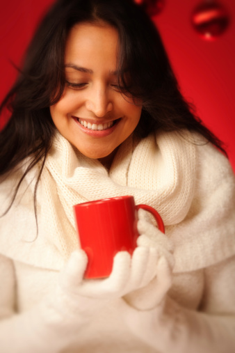 Subject: Vertical close-up view of a smiling, young, pretty hispanic woman dressed in warm, white winter clothing and holding a warm drink in a red mug for the cold winter season. The red background includes hanging Christmas balls for the holiday season.