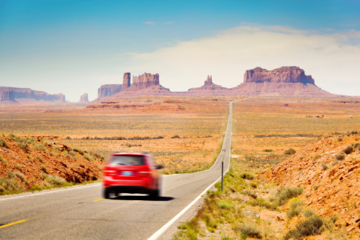 Subject: Tourists in a red car traveling in the American Southwest, driving downhill on a straight length of highway stretching from southern Utah toward Monument Valley, Arizona