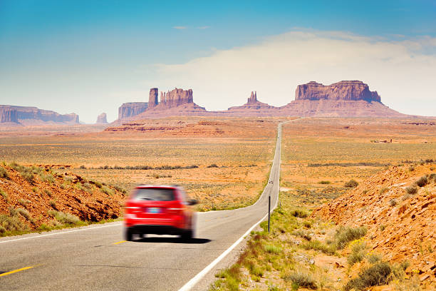 Car Tourist Traveling in the American Southwest, Monument Valley, Arizona Subject: Tourists in a red car traveling in the American Southwest, driving downhill on a straight length of highway stretching from southern Utah toward Monument Valley, Arizona the mittens monument valley stock pictures, royalty-free photos & images