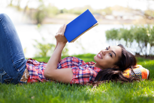 Beautiful mixed race young student laying down in a field while reading a book.